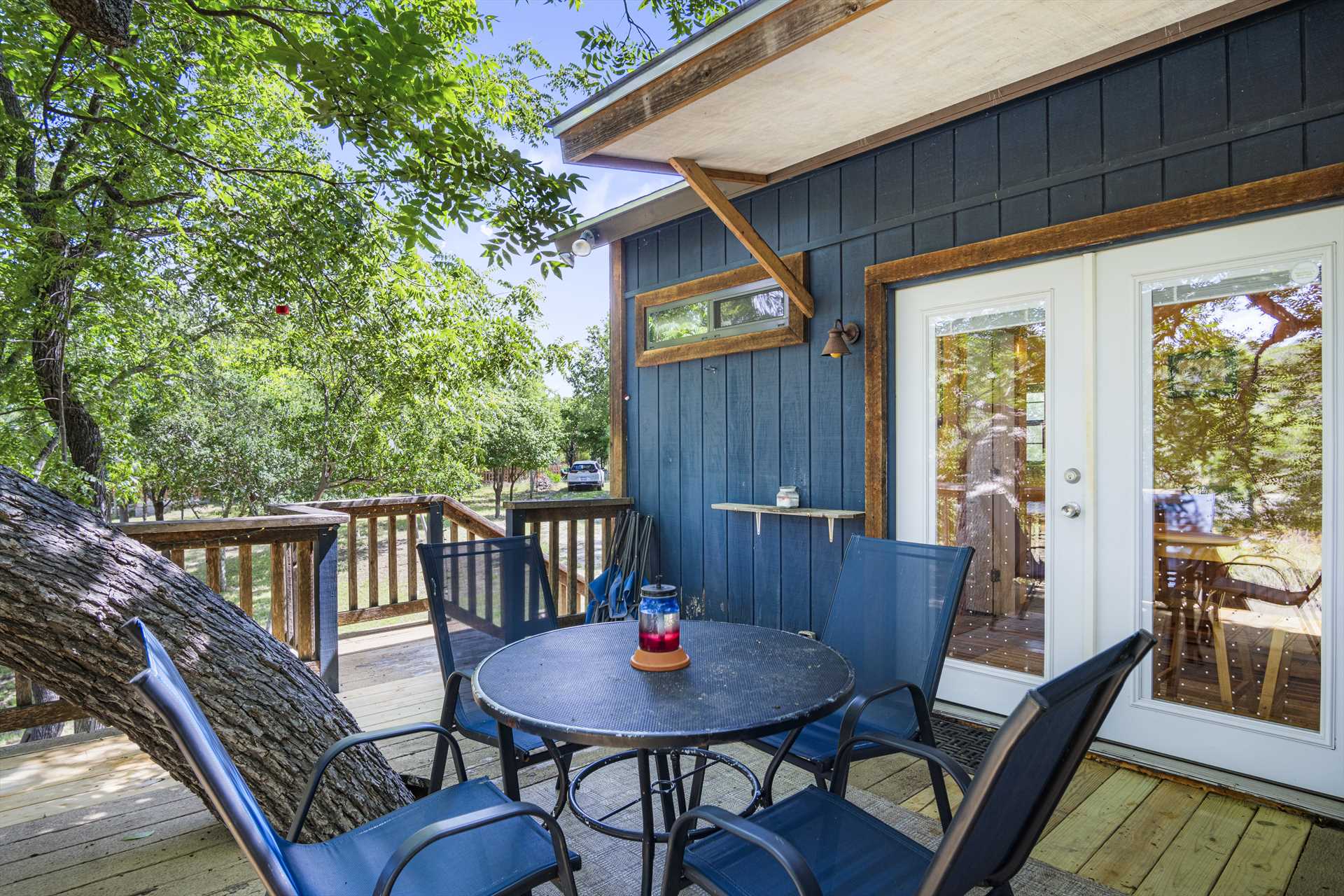 Outdoor seating area featuring a round table and chairs on a deck, surrounded by greenery and a blue siding house, ideal for relaxation.