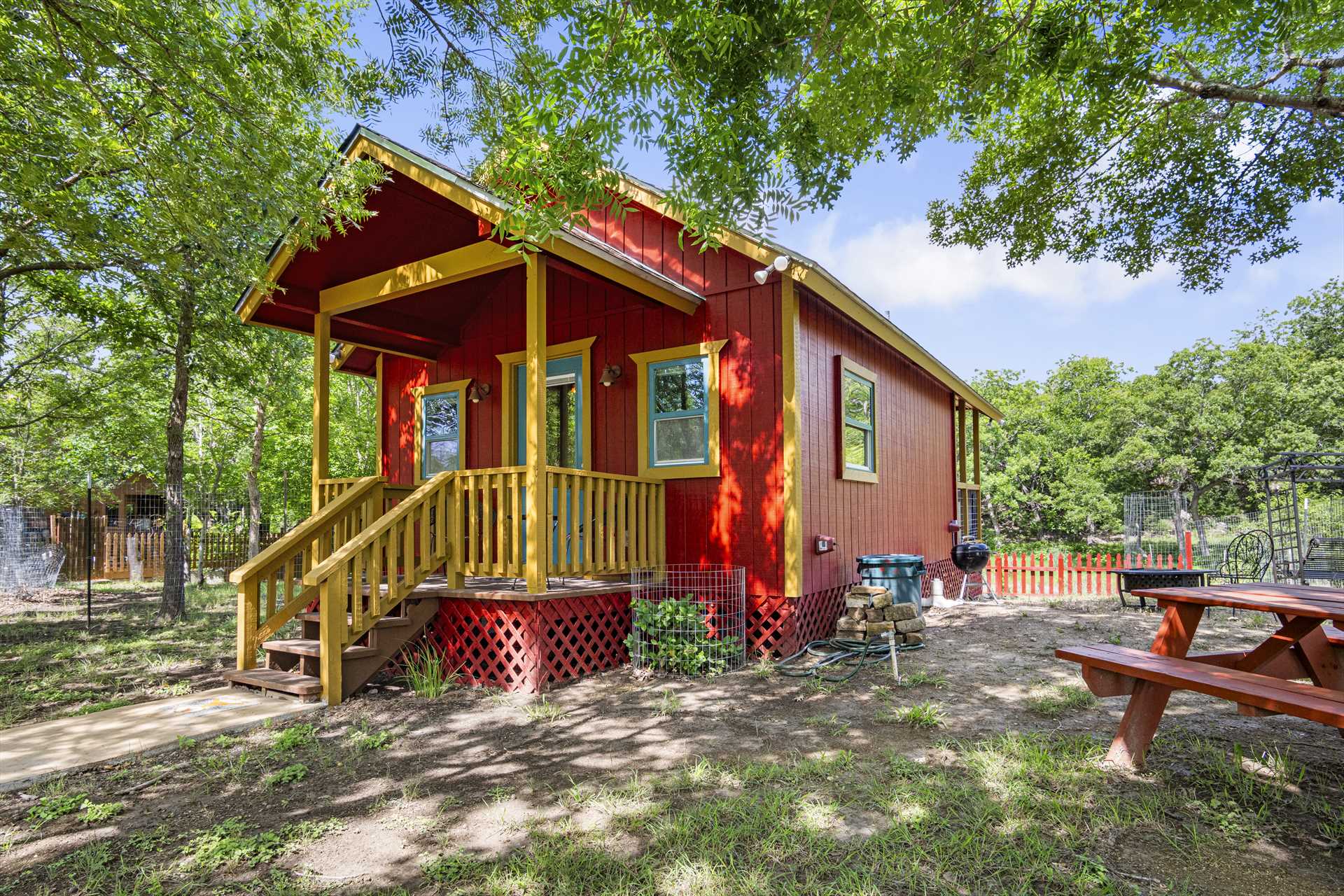 Colorful red cabin with yellow trim, wooden steps, and a picnic table, nestled among green trees, showcasing a cozy outdoor space.