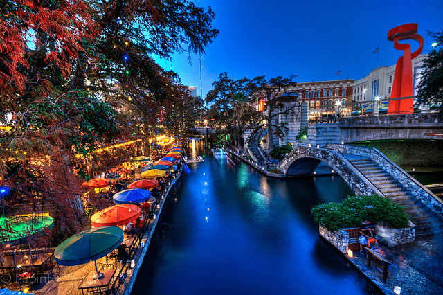 Twilight view of a vibrant riverwalk lined with colorful umbrellas and dining spots, showcasing lights and reflections along the water.