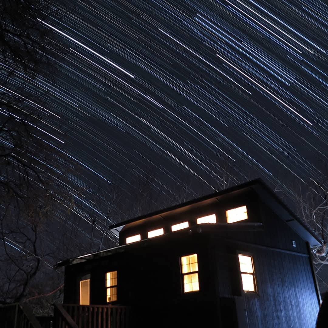 Star trails illuminate a dark sky above a cozy, lit cabin nestled among trees, showcasing the beauty of night photography and celestial movement.