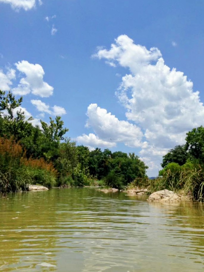 Serene river scene with lush greenery along the banks, under a bright blue sky filled with fluffy clouds. Ideal for nature exploration.
