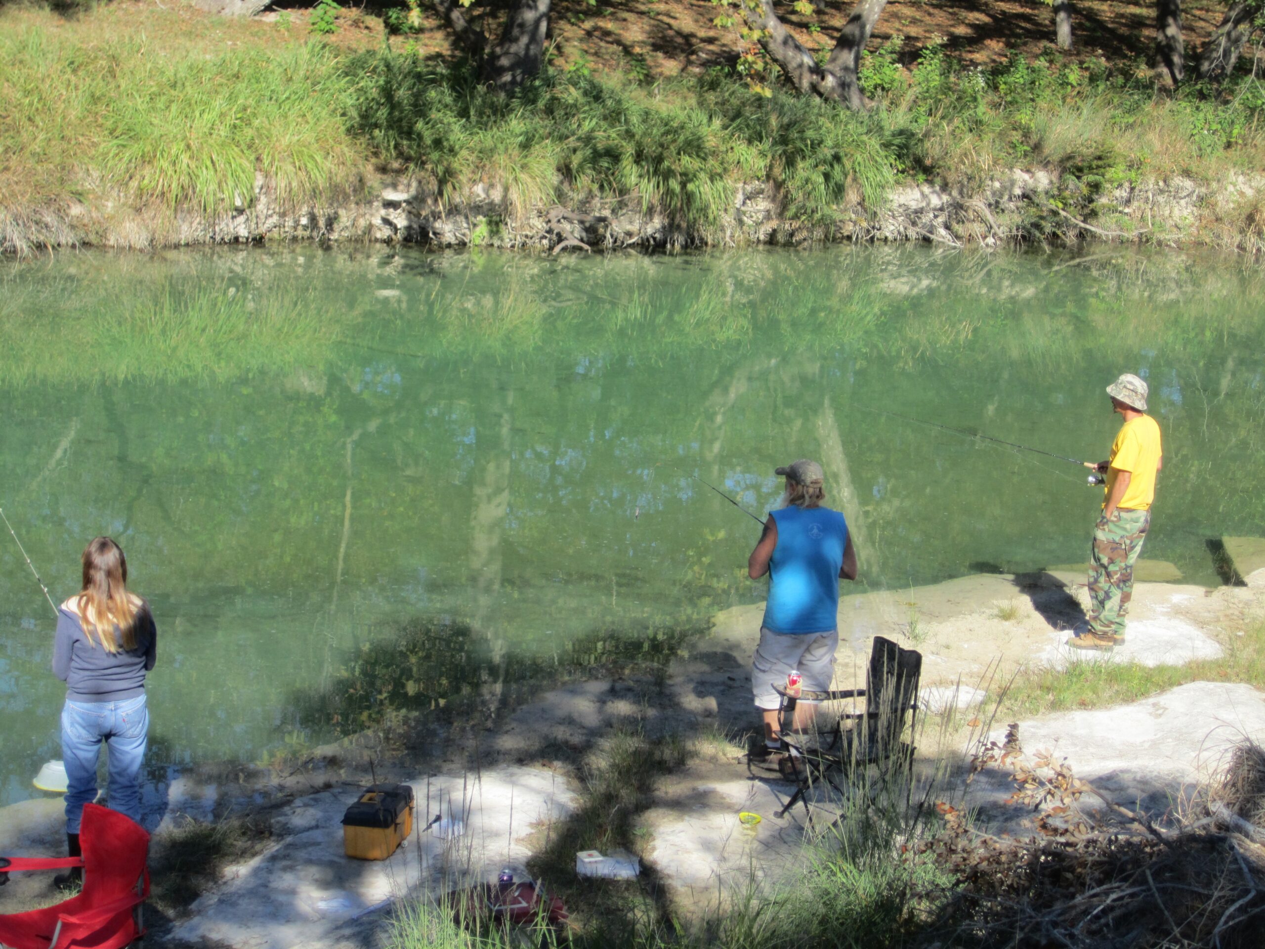 Three people fishing by a tranquil river, surrounded by greenery. The scene captures a peaceful outdoor activity, highlighting relaxation and nature.